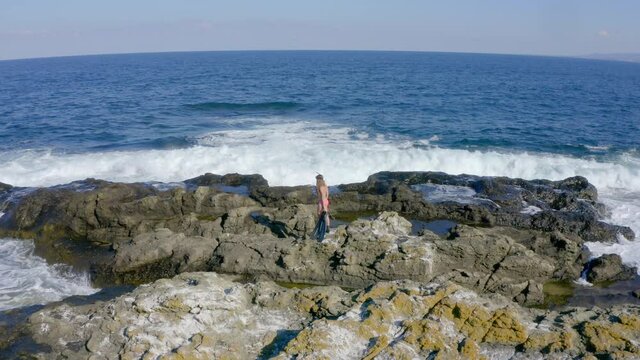 Drone shot of a young woman over the sea rocks. Seafoam spills over the brown rocks and everything is surrounded by the choppy blue sea.
