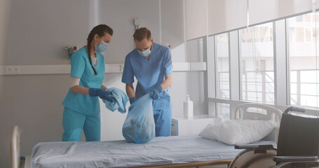 Nurses in safety mask and gloves making the bed and changing sheets in hospital ward