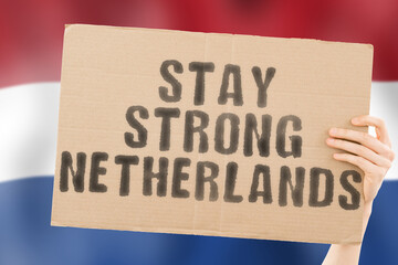 The phrase " Stay strong Netherlands " on a banner in men's hand with blurred Dutch flag on the background. Support. Patriot. Community. Free. Freedom. Independence. Nation. Problem. Issue