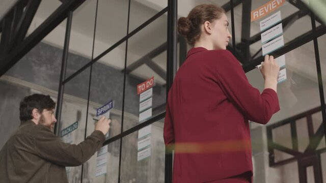 Low angle rear of two Caucasian business woman and man getting ready for corporate presentation, working with note cards on glass wall, writing