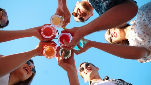 Low angle view of friends having fun at pool party, clinking glasses with colorful summer cocktails near hotel swimming pool. People toast drinking fresh juice at luxury summer villa in slow motion.