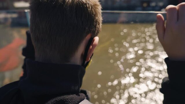 A Teenager Looks Through The Glass Down At The Water Standing On The Embankment In The City. Shooting From Behind