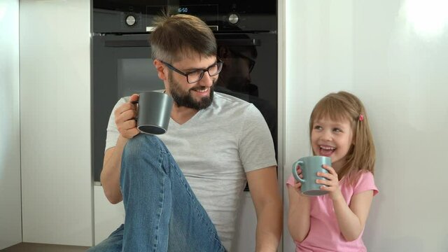 Happy family, smiling father and little daughter sit on floor in kitchen and drink coffee from cups. Father and child girl have rest, laugh and fun talk to each other. Leisure