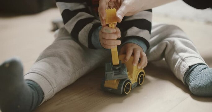 View of baby boy playing excavator on the wooden floor. Mothers hand shows him how to dig. Room filled with morning sunlight. Tiny fingers with toys.