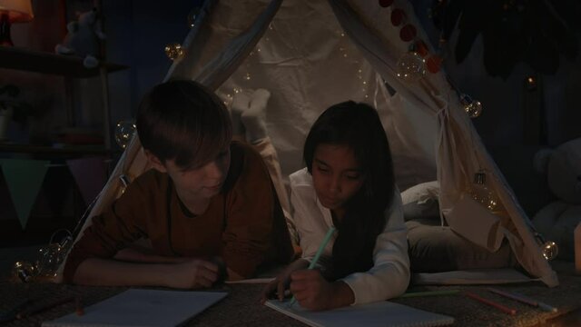 Young boy and girl talking and creating pictures while spending free time in evening. Teenagers lying on floor and talking in decorative makeshift hut while drawing with colored pencils.