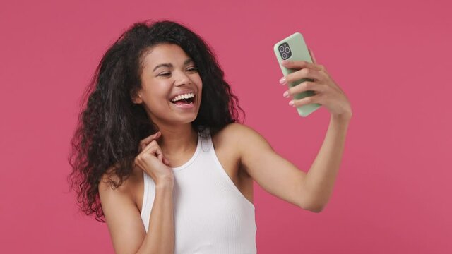 Young african woman curly hair 20s years old wears white tank top shirt doing selfie shot on mobile phone post photo on social network swipe isolated on dark pink color wall background studio portrait