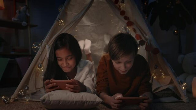 Positive teenagers playing online games in decorative makeshift hut at home in evening. Girl and boy lying on floor having fun while using smartphones.Concept of leisure.