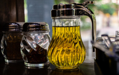 Close up shot of olive oil kept along with chilli, pepper sprinkler and blurry glass window in the background.