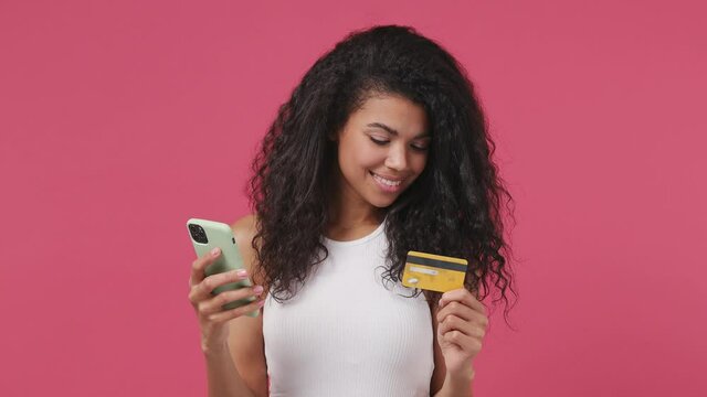 Young woman wear white tank top shirt using mobile cell phone hold in hand credit bank card doing online shopping order delivery to home booking tour isolated on dark pink color wall background studio