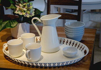 Dishes for the evening dinner. On a platter are cups, a milk jug and a gravy boat, theres a stack of bowls in the shade nearby. A side light illuminates the table.