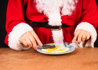 Christmas healthy breakfast of Santa. Close up of Santa Claus's hands in traditional red costume with knife and fork slicing scrambled eggs.