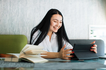 Happy joyful likable Asian woman uses digital tablet, while sitting at the table in coffee shop....