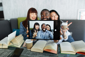 Group of three young multiethnic friends, sitting at the table in cafe, reading books, taking selfie using digital tablet pc, laughing, gesturing. Focus on tablet computer