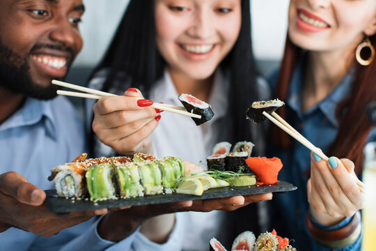 Lunch at a Asian restaurant. Close up shot of happy grinning multiethnic friends, black man and two pretty girls, eating sushi rolls with chinese sticks. Focus on plate with rolls and sticks