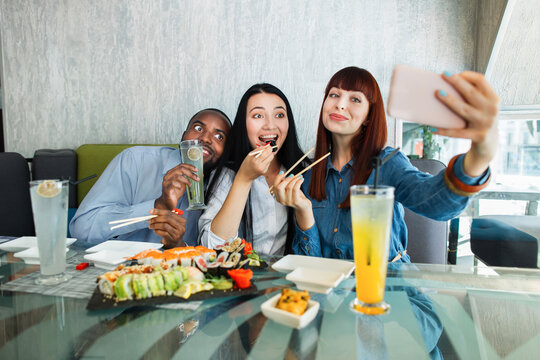 Selfie time, lunch and friendship concept. Pretty red haired girl taking a picture by the cell phone with her friends, Asian woman and African man, enjoying sushi food