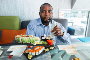 Wow, that is cool and tasty. Excited funny African handsome man, drinking cold fresh juice cocktail in cafe and holding big plate served with delicious sushi rolls