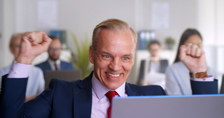 Happy mature businessman working on laptop making winner gesture celebrating success.