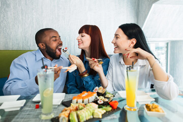 Happy three multiethnic friends having fun at cafe eating chinese food and sushi rolls. Pretty funny Caucasian girl feeding her male laughing African friend with sushi roll on wooden stick