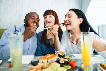 Young jolly multiracial friends eating sushi using chopsticks, looking positive and happy, sitting...