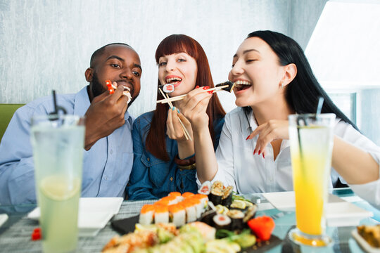 Young Jolly Multiracial Friends Eating Sushi Using Chopsticks, Looking Positive And Happy, Sitting At Asian Restaurant And Smiling. People, Emotions, Food And Friendship Concept