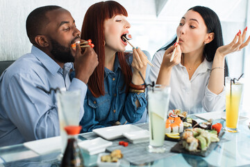Lunch at a Asian restaurant. Close up shot of happy funny multiethnic friends, black man and two pretty girls, eating sushi rolls with chinese sticks
