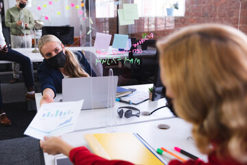 Caucasian woman wearing face mask passing a document to her female office colleague at office