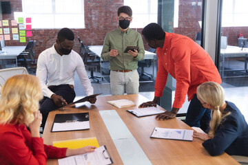 Team of diverse male and female office colleagues wearing face masks discussing together at office