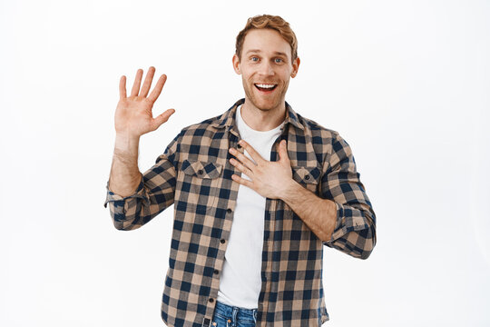Image of smiling redhead man holding one raised and another hand on heart, introduce himself, my name is nice to meet you gesture, saying hello, standing friendly and happy against white background