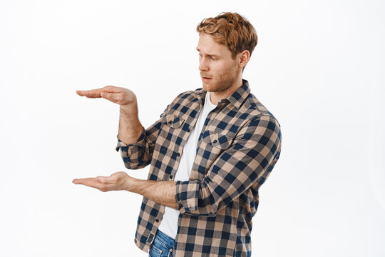 Image of adult man with red hair and beard, holding something empty space, looking at hands displaying an item with confused face, standing over white background