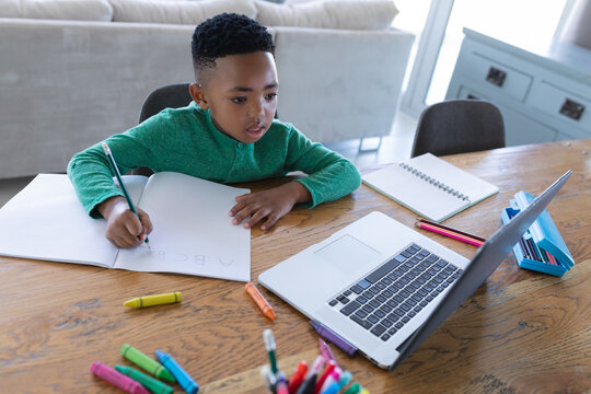 African american boy in online school class, using laptop and writing in his notebook