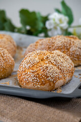 Fresh challah bread rolls hot from the oven on baking tray. Gluten free bread buns or mini loaves with sesame seeds and golden color ready to eat. Fluffy shaped homemade bread, flowers in background.