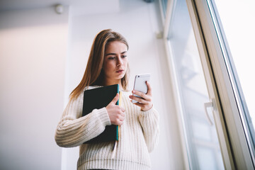 Serious young woman standing with textbooks and using smartphone