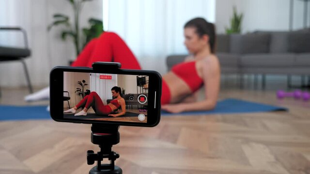 Fit woman fitness trainer blogger in sportswear tells teaches student doing workout exercise gluteal bridge while lying on yoga mat. Sports girl records online course lesson on smartphone in home