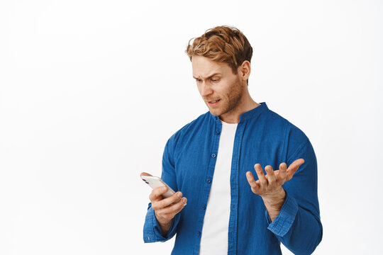 Image of confused and annoyed young man looking at smartphone, complaining on application, read bothering ridiculous message, raising hand irritated, standing over white background