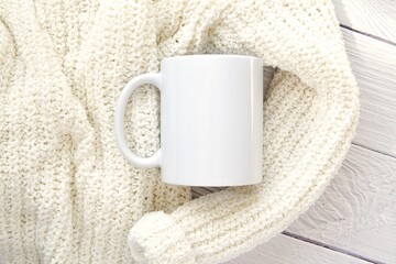 White ceramic coffee mug mockup for design presentation, soft woman sweater on washed wood background, aesthetic composition.