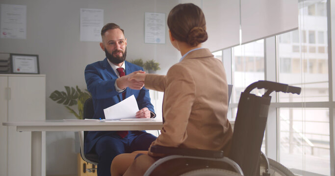 Young woman in wheelchair talking with employer and shaking hands in office