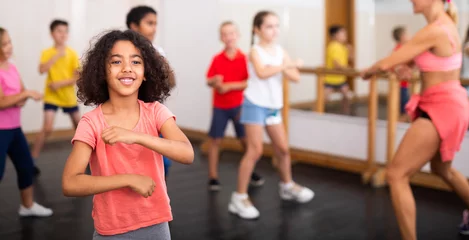 Photo sur Plexiglas École de danse Portrait of cheerful little African American girl training movements during group class in dance school