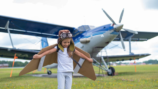 A little girl in a pilot's costume with cardboard wings runs on the lawn against the backdrop of the plane. A child in a hat and glasses dreams of flying on an airplane.