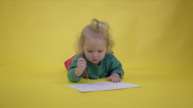 Barefoot adorable happy toddler girl wearing green draws with brush on sheet of paper on yellow studio background copy space. cute child artist creating children's art. Concept of early development
