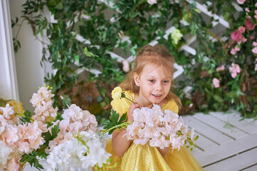 Obraz na płótnie Canvas Girl, 7, sits against a green wall with flowers in a yellow dress and sniffs a white flower