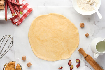 Home baking concept with raw dough rolled out with ingredients flour, water, butter, sugar and rhubarb for baking on white marble background. Top view of homemade pie crust on the table.