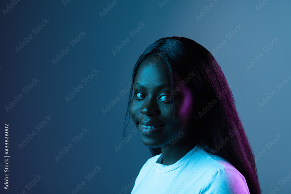 Wall mural Portrait of young African beautiful girl isolated over dark background in mixed neon light.