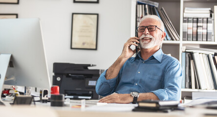 Senior business manager sitting at the desk in his office.He speaks on smart phone.	