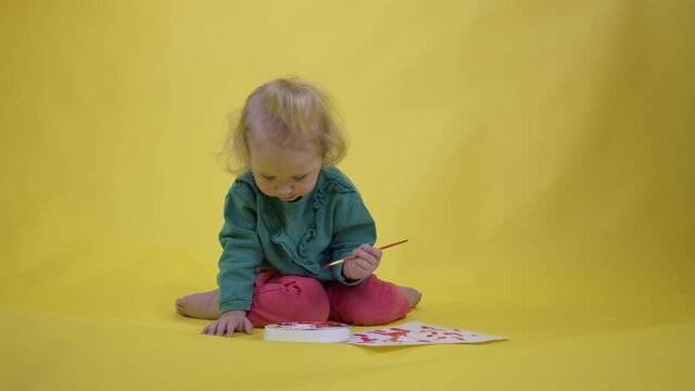 adorable happy toddler girl wearing green Picks up paint on palette and draws child's picture on yellow studio background copy space. cute child artist creating children's art.