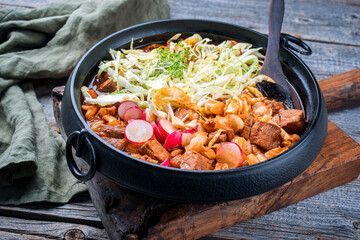 Traditional slow cooked Mexican pozole rojo soup with pork meat and hominy maiz served as close-up in a modern design cast-iron roasting dish on an old rustic wooden board