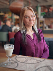 A blonde girl in a lilac sweater sits in a cozy restaurant on a sofa and she is relaxed and smiling and have a coffee drink on her table.