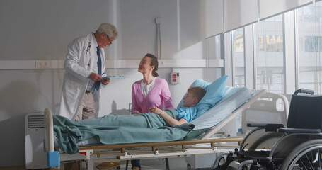 Mature doctor talking to young mother and sick son lying in hospital bed