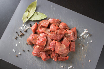 Raw beef of veal cut into cubes on a gray cutting board next to bay leaves, black pepper peas and coarse salt. Ingredients for making a delicious home-made meal