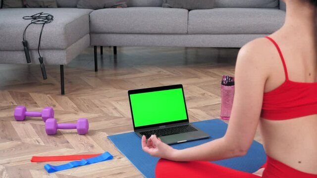 Green screen mock up chroma key monitor laptop: Calm sportive woman in sportswear practices yoga online video call webcam computer at home. Sports girl sits meditating in lotus position on fitness mat