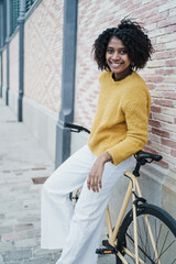 Fototapeta na wymiar Portrait of cheerful carefree black woman with afro hairstyle smiling and standing next to cool yellow bike and pink brick wall